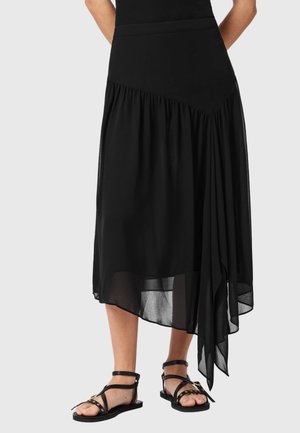 Jupe midi asymétrique noire avec détails froncés, portée avec des sandales plates noires ornées de clous dorés et de brides à la cheville.