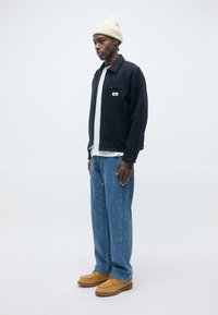 Joven con un gorro blanco, chaqueta navy, camiseta blanca, jeans azules y zapatos color beige, de pie frente a un fondo blanco liso.
