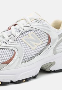 Białe i szare buty sportowe z siateczkowymi bokami, beżowymi i różowymi akcentami, białymi sznurowadłami oraz dużym logo "N" po boku.