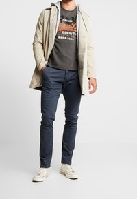 Světlý béžový trenčkot přes šedou mikinu s kapucí, černé tričko s potiskem, tmavě modré úzké kalhoty a krémové tenisky. Jednoduchý design s kombinací barev.