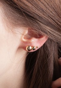Złote kolczyki-studki z trzema kulistymi elementami o różnych rozmiarach, gładkiej fakturze i wypolerowanym wykończeniu, noszone na małżowinie usznej.