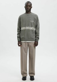 Pelēks adīts džemperis ar tekstuālām detaļām, apaļu kakla izgriezumu un kontrastējošām aprocēm, kopā ar bēšām biksēm un melniem apaviem. Vienmērīga tekstūra kopumā.