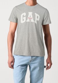 T-shirt en coton gris arborant "GAP" en lettres blanches avec "Original" en rouge en dessous. Manches courtes, col rond, coupe décontractée.