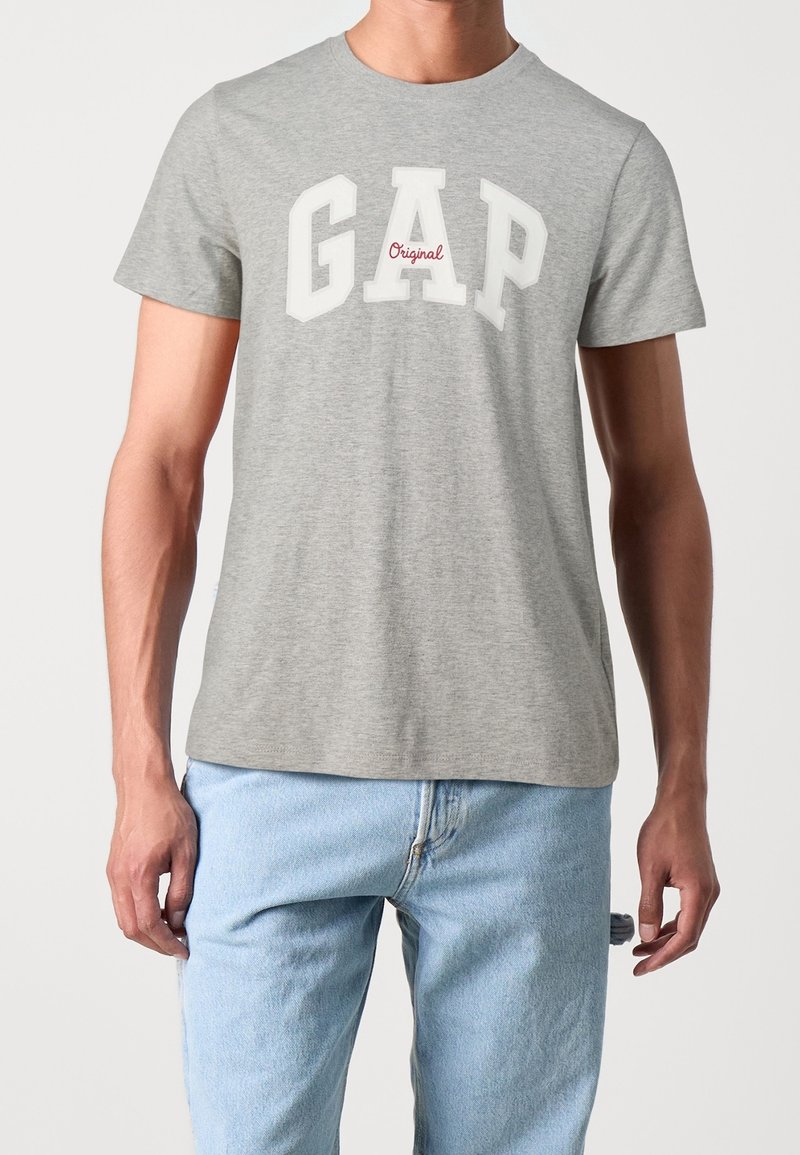T-shirt en coton gris arborant "GAP" en lettres blanches avec "Original" en rouge en dessous. Manches courtes, col rond, coupe décontractée.