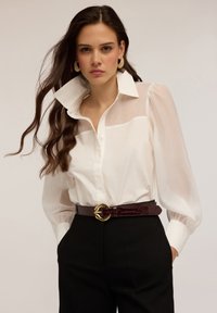 Blouse blanche avec des manches transparentes et un col structuré, associée à un pantalon noir taille haute et une ceinture bordeaux avec une boucle dorée.