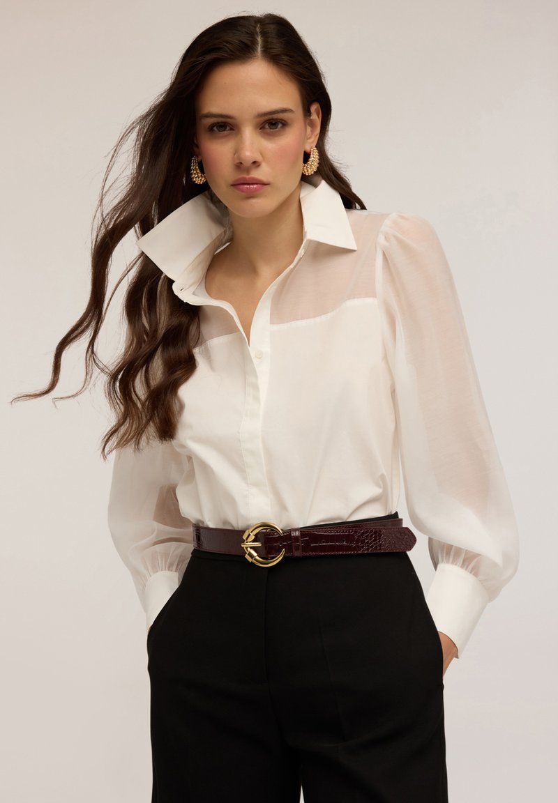 Blouse blanche avec des manches transparentes et un col structuré, associée à un pantalon noir taille haute et une ceinture bordeaux avec une boucle dorée.
