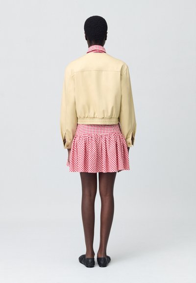 Veste bomber beige avec ourlet élastique et manches longues sur une mini-jupe vichy rouge et blanche à volants. Des chaussures noires complètent le look.