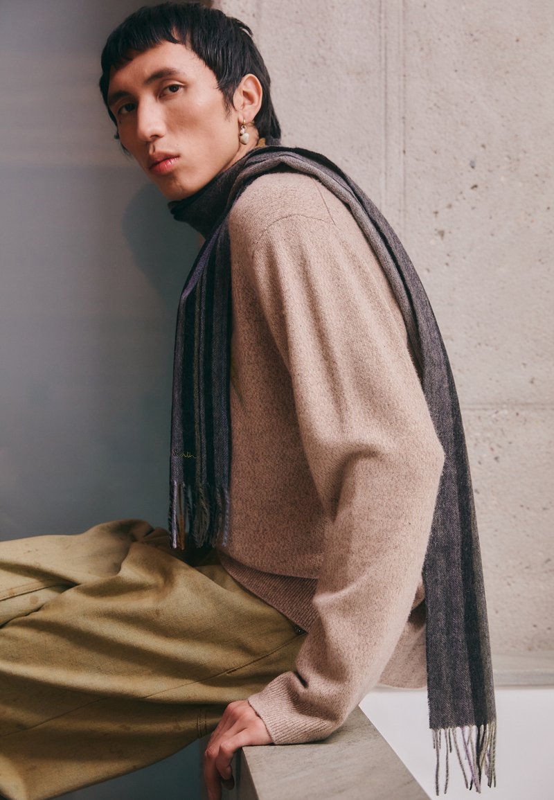 Pull en maille beige avec un col montant, associé à un pantalon large vert olive. Une écharpe rayée gris foncé repose sur l'épaule.