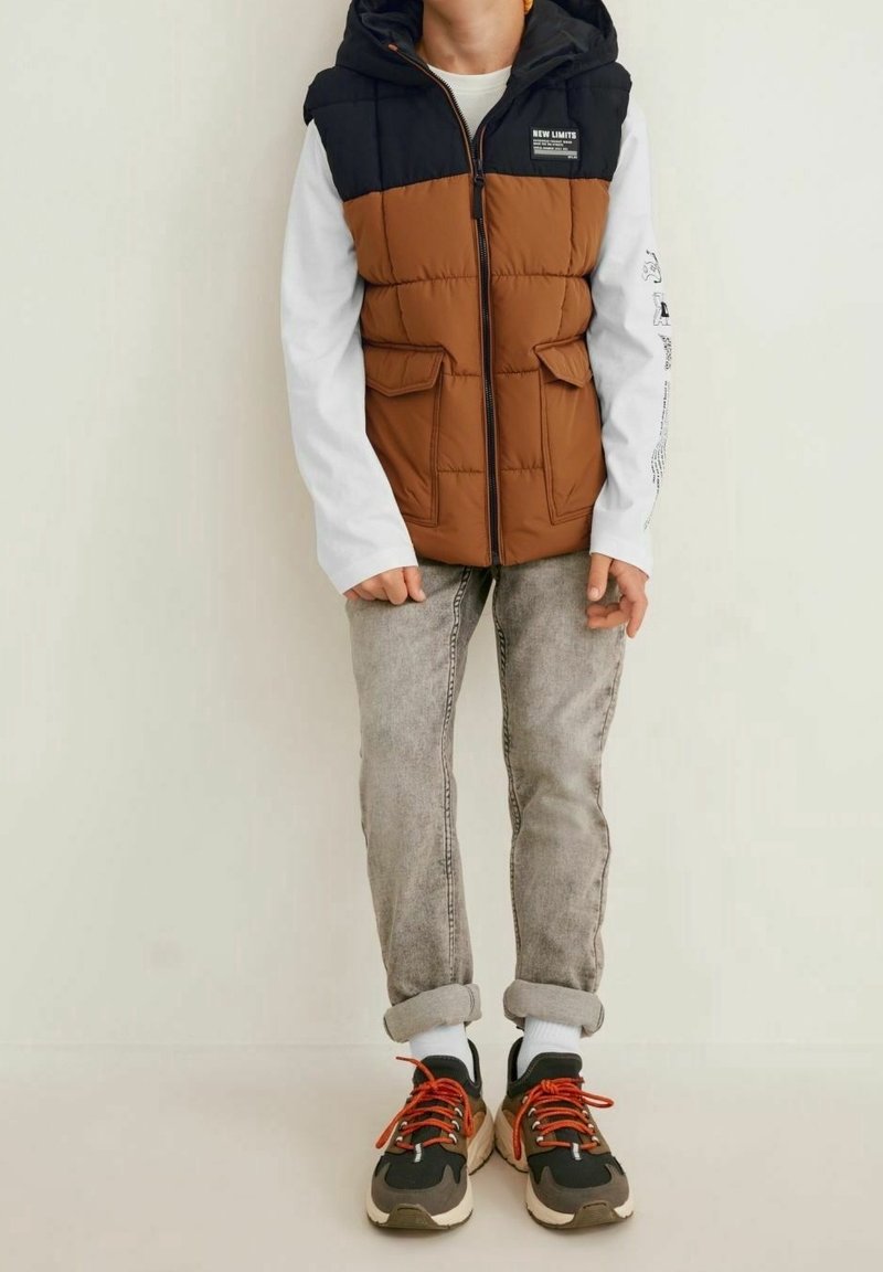 Puchowa kamizelka w kolorze brązowym i czarnym z kapturem, przednimi kieszeniami i zamkiem; zestawiona z szarymi dżinsami i czarnymi trampkami z pomarańczowymi sznurowadłami.