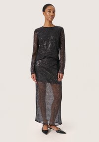 Abito lungo nero a maniche lunghe con sovrapposizione trasparente e paillettes; corpino aderente con gonna fluida; abbinato a ballerine nere con punta affilata.