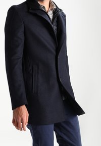 Hombre vestido con un abrigo marino oscuro sobre una camisa con estampado y pantalones azules, frente a un fondo blanco liso.