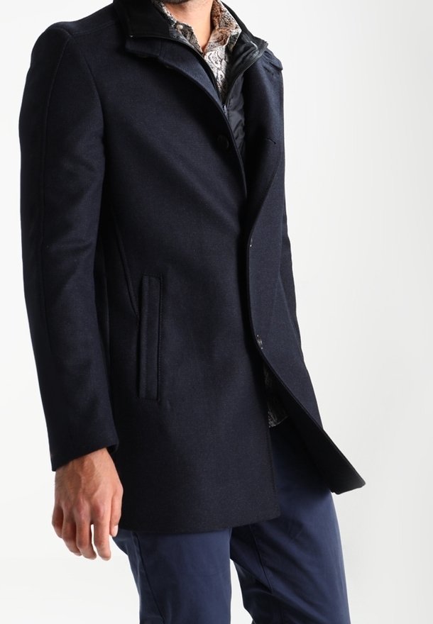 Hombre vestido con un abrigo marino oscuro sobre una camisa con estampado y pantalones azules, frente a un fondo blanco liso.