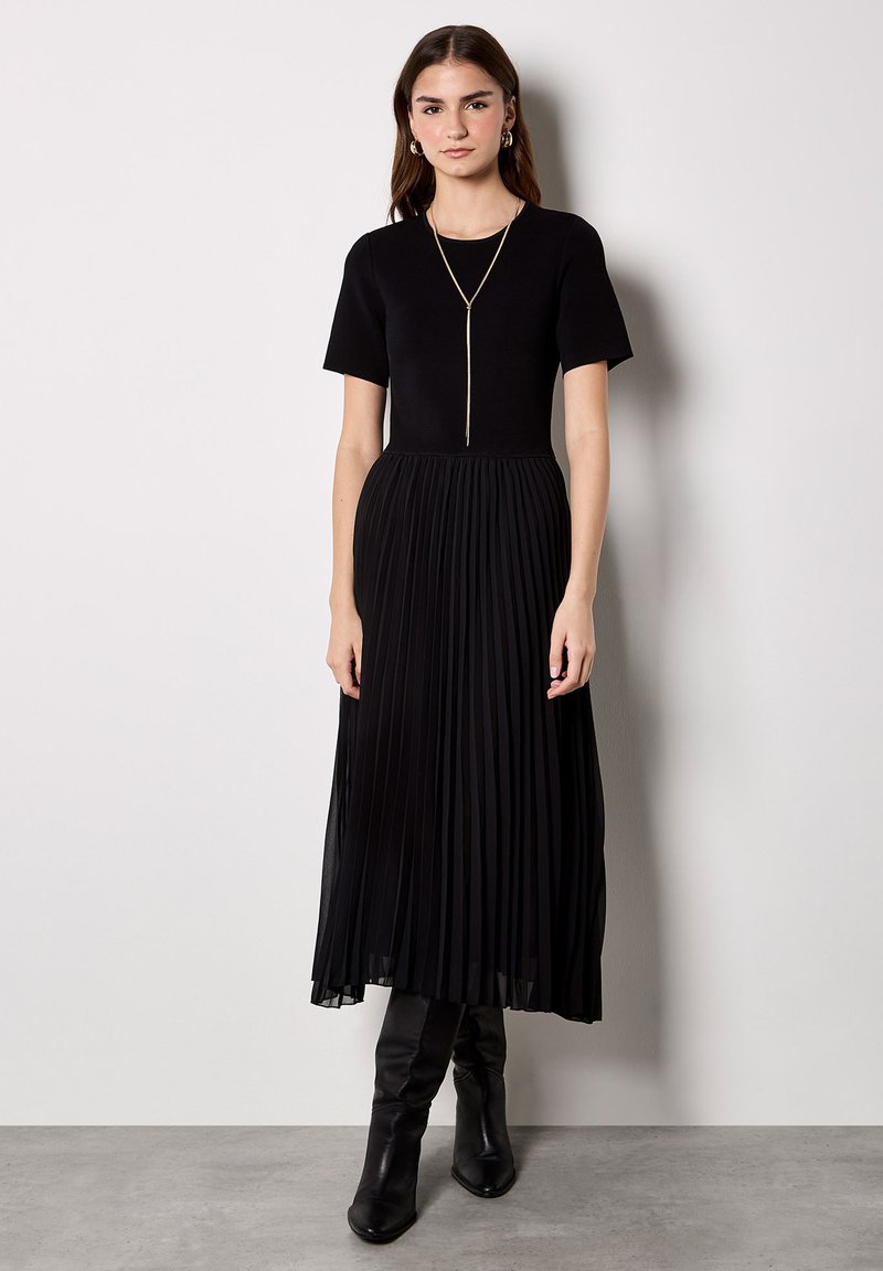 Robe noire plissée à manches courtes, avec un corsage ajusté et une jupe fluide. Portée avec des bottes noires montantes jusqu'au genou et un collier en or.