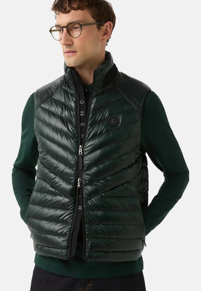 Mann trägt eine dunkelgrüne, gesteppte, ärmellose Jacke über einem grünen Langarmhemd, die Hände in den Taschen, und schaut leicht nach links.