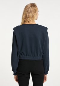 Felpa corta blu navy con vita elastica, maniche lunghe e ampie, e dettagli sulle spalle. Texture liscia, design con collo tondo.