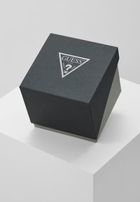 Black textured box with a triangular shape, featuring a silver GUESS logo on top and a contrasting gray base.