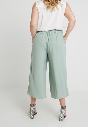 Vrouw staat met haar rug naar voren gekeerd, draagt een witte mouwloze blouse, lichtgroene wijde cropped pants en sandalen met hoge hak en slangenprint.