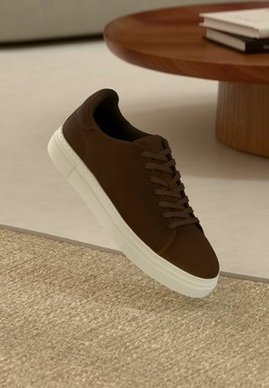 Brūna zamšāda sneaker ar plakanām auklām un baltu gumijas zoli, ar noapaļotu purngalu un minimālistisku dizainu, novietota uz teksturētas virsmas.