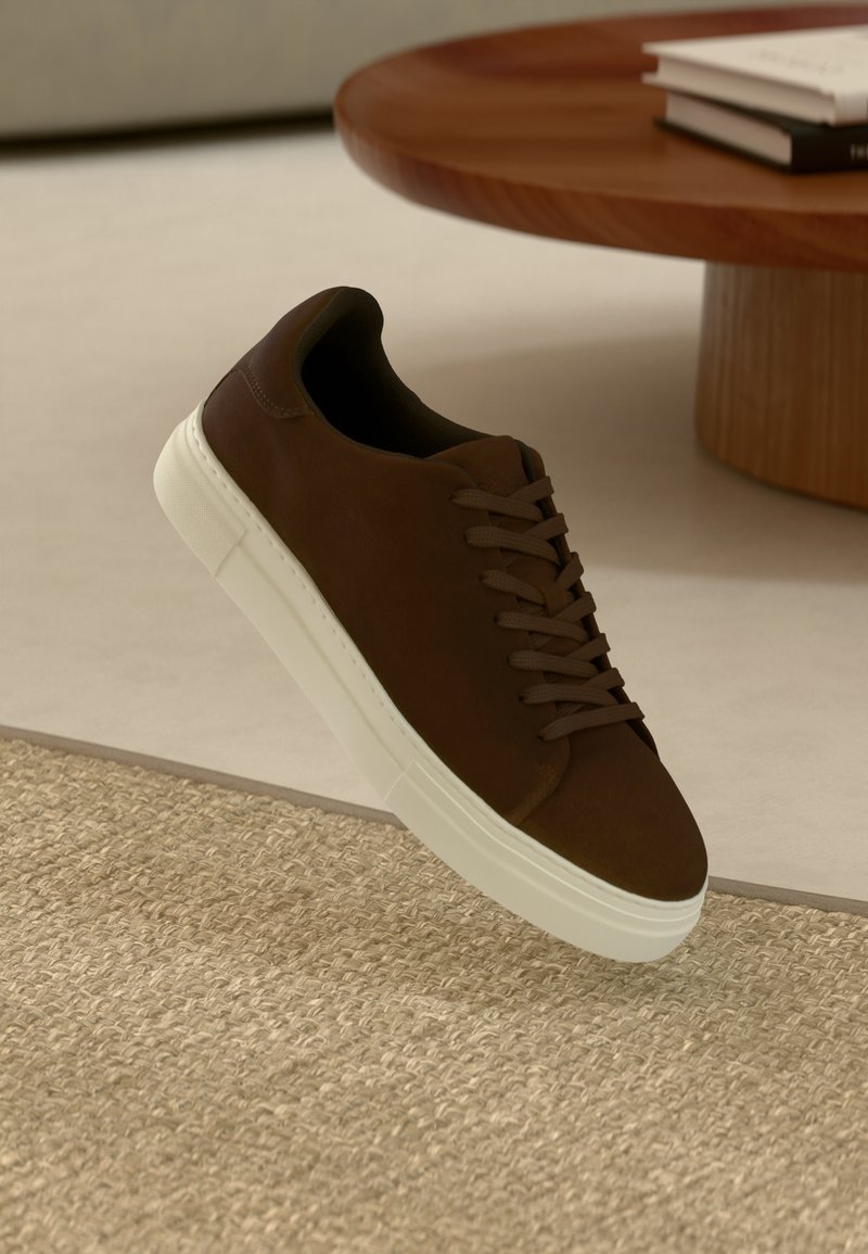 Sneaker in suede marrone con lacci piatti e suola in gomma bianca, caratterizzato da una punta arrotondata e un design minimale, posizionato su una superficie texturizzata.