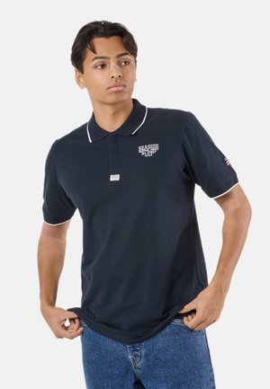 Giovane uomo che indossa una polo blu navy con rifiniture bianche e scritta sul petto, guarda di lato, tiene l’orlo della maglia, in piedi su uno sfondo semplice.