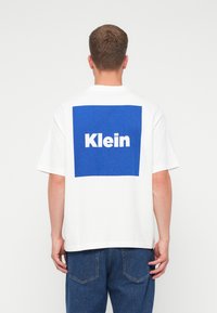 Mężczyzna ubrany w biały T-shirt z dużym niebieskim kwadratem i napisem "Klein" na plecach, w połączeniu z niebieskimi dżinsami, stojący na tle białym.