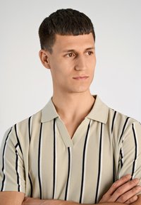 Beżowa koszulka polo z pionowymi granatowymi paskami, teksturowana tkanina i dekolt w kształcie V. Model pozuje z rękami skrzyżowanymi, na neutralnym tle.