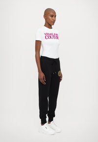 Hvit tettsittende t-skjorte med rosa "VERSACE JEANS COUTURE"-tekst, svarte joggebukser med et gulllogo-lapp, og hvite sneakers. Enkelt design, avslappet passform.