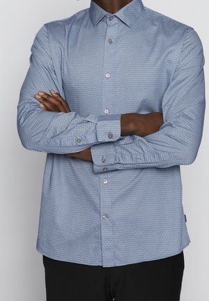 Homme portant une chemise bleu clair à motifs avec boutons et manches longues, debout les bras croisés devant un fond neutre.