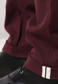 Bordeauxrode stoffen broek met taps toelopend ontwerp en een wit label met een zwarte streep aan de zoom, gecombineerd met zwarte schoenen.