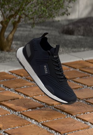 Black athletic shoe with textured fabric upper, white sole, and laces. Features logo and synthetic accents, suspended above a patterned surface.