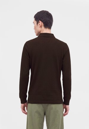 Homme de dos portant un polo à manches longues marron foncé et un pantalon vert clair sur un fond clair uni.