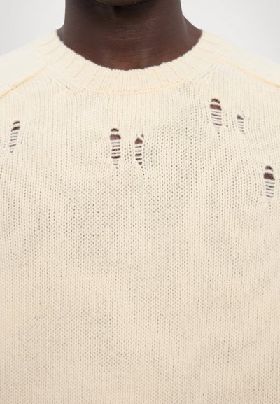 Pulover din tricot bej deschis, cu o guler cu aspect texturat și mai multe găuri deteriorate în întreaga țesătură, evidențiind un design texturat, uzat.