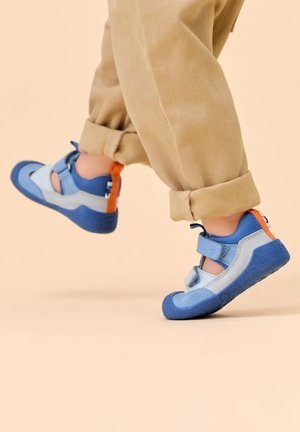 Kind trägt blaue und graue Sneakers mit orangefarbenen Akzenten, hebt die Füße mitten im Schritt, beigefarbene Hose über den Knöcheln hochgekrempelt auf beigem Hintergrund.