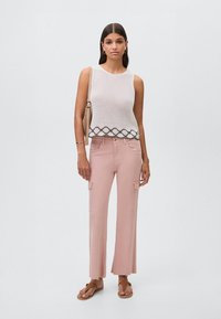 Bílý pletený top bez rukávů s vlnkovaným lemem, doplněný světle růžovými širokými cargo kalhotami, které mají kapsy a roztrhané okraje. Ploché sandály.