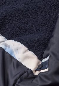 Un tissu en polaire bleu marine complété par une doublure bleue plus claire et lisse. Il présente un bouton et une couture contrastante le long du bord.