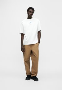 Λευκό κοντομάνικο μπλουζάκι με μπλε λογότυπο στο στήθος, συνδυασμένο με άνετο μπεζ παντελόνι και μαύρα παπούτσια.