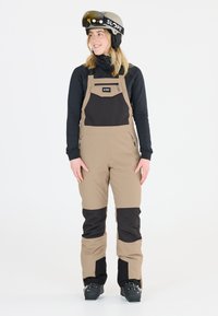 Beige und schwarze Skihose mit einer Reißverschlusstasche auf der Brust, aus wasserabweisendem Material. Getragen mit einem schwarzen Langarmshirt und Helm.