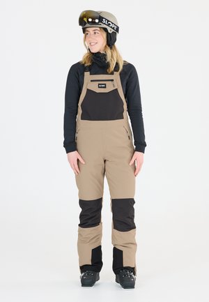 Beige en zwarte skibroek met een ritszak op de borst, gemaakt van waterafstotend materiaal. Draagt een zwarte lange-mouwen top en een helm.