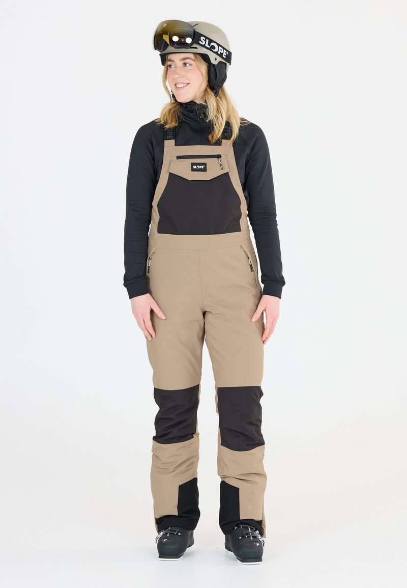 Beige und schwarze Skihose mit einer Reißverschlusstasche auf der Brust, aus wasserabweisendem Material. Getragen mit einem schwarzen Langarmshirt und Helm.