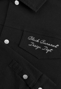 Zwarte shirt met een textuurstof. Beschikt over zilveren knopen en een rechthoekige patch met "Black Bananas Design Dept." geborduurd in het wit.
