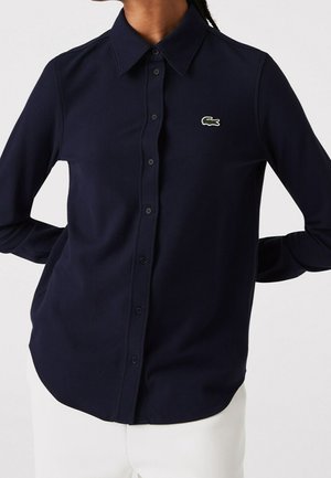 Osoba ubrana w granatową koszulę na guziki z długim rękawem, z małym zielonym logo krokodyla na lewej piersi, w połączeniu z białymi spodniami.