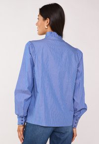 Camicia a righe blu e bianche con colletto alto, maniche a sbuffo e polsini con bottoni. Realizzata in un tessuto leggero con una texture liscia.