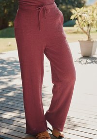 Bordeauxfarbene weit geschnittene Hose aus strukturiertem Stoff. Sie verfügt über einen Kordelzug am Bund und einen lockeren Schnitt, kombiniert mit flachen Sandalen auf einer Holzoberfläche.
