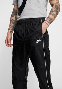 Melni neilona sporta bikses ar elastīgu jostasvietu, sānu kabatām, baltām apmalēm un mazu baltu Nike logo kreisajā augšstilbā. Gluda tekstūra.