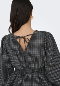 Femme aux longs cheveux foncés portant une robe à carreaux noire et grise avec des manches longues bouffantes et un détail noué au dos de l'encolure.