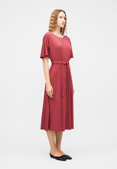 Γυναίκα με κυματιστά μαλλιά που φοράει ρόζ φόρεμα μέχρι τα γόνατα με κοντά μανίκια, ζώνη δεμένη στη μέση και μαύρες μπαλαρίνες, όρθια πλάγια.