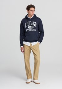 Młody mężczyzna w granatowej bluzie z kapturem z napisem "Chicago 1983 Athletic", białej koszulce pod spodem, beżowych spodniach i białych trampkach, stojący na jednolitym tle.