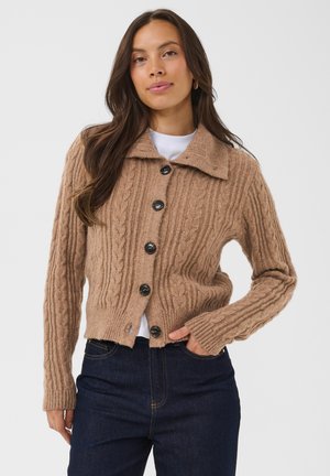 Femme aux longs cheveux bruns portant un cardigan boutonné beige en maille torsadée et un jean foncé, debout avec une main dans la poche devant un fond uni.