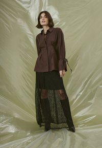 Brown button-up shirt with side ties, paired with a black lace maxi skirt featuring sheer panels and a textured pattern.