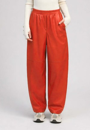 Personne portant un pantalon large rouge vif, un haut blanc à manches longues et des baskets blanches, debout avec une main dans la poche.